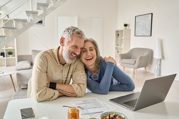  sitting at home table with laptop. Smiling middle aged senior 50s husband and wife having fun satisfied with buying insurance, paying bills online.
