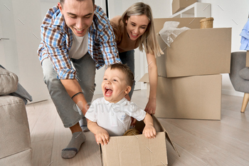 Happy parents playing with cute toddler kid son riding in box on moving day. / Photodune