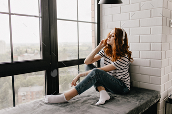 Cute ginger lady in jeans sitting on sill and listening music. Pleased curly girl posing beside win / Photodune