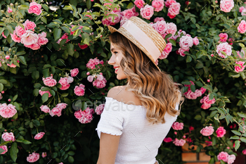 Lovely blonde girl in summer hat looking at flowers with smile. Pleased curly woman relaxing during / Photodune