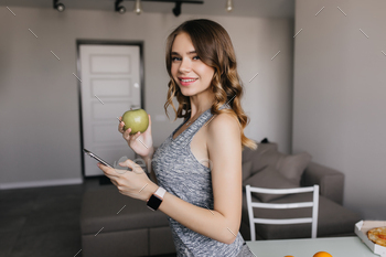 Slim pale girl in smartwatch posing at home. Charming dark-haired lady with apple having fun in mor / Photodune