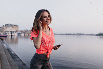 Good-humoured girl listening music after training. Attractive female runner posing near river with / Photodune