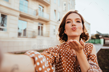 Blissful girl in vintage attire making selfie on the street. Gorgeous caucasian lady in brown outfi / Photodune