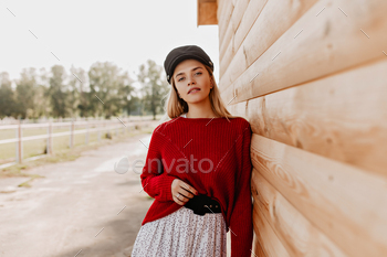 Pretty young blonde in red sweater and dark hat posing in park near wooden wall. Beautiful woman we / Photodune