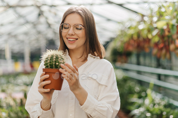 Smiling young woman touching cactus. Photo of joyful lady with plant posing in orangery / Photodune