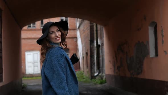 Young Redhead Woman in a Blue Coat and Black Hat with a Suitcase on the Background of the Old City