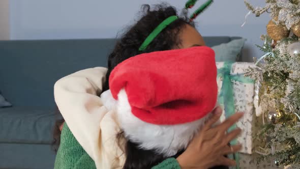 Little Girl in a Christmas Hat Hugs Her Mother Against the Background of a Festive Fir Tree and