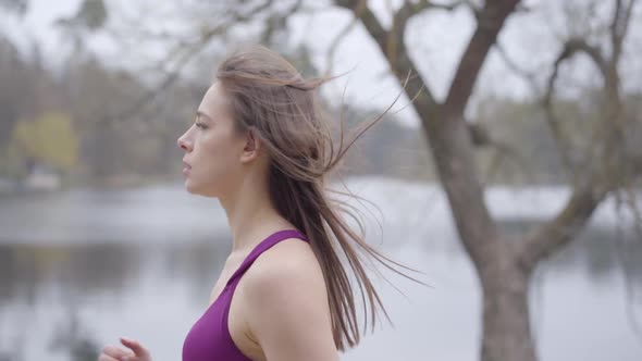 Side View of Confident Caucasian Sportswoman Running Along Lake Shore in Park