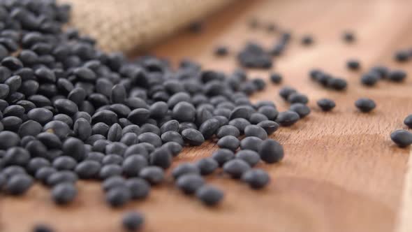 Dark black lentil seeds fall onto a burlap wooden surface in slow motion