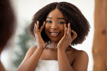 cking first wrinkles, touching her skin after applying cream at home. African American woman looking at her reflection after skincare procedures