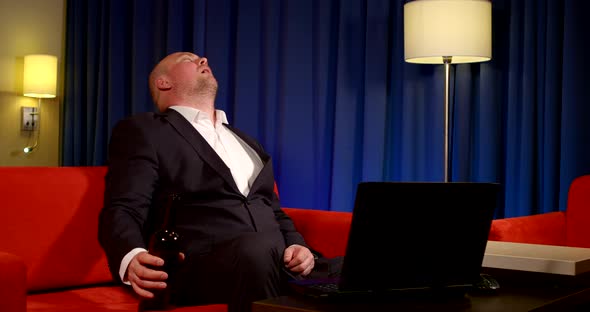 a Bald Man in a Dark Suit Is Sitting on a Red Sofa in Front of a Laptop Against a Blue Curtain