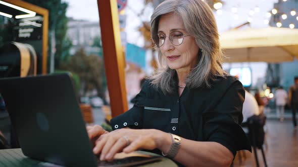 Stylish Adult Grayhaired Businesslady in Glasses is Attentively Writing a Message on Her Laptop