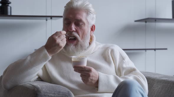 A Handsome Elderly Man with a Gray Beard Eats Yogurt Holding a Spoon Close To His Mouth