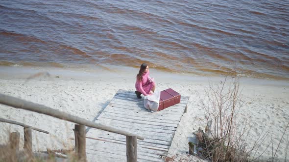 Wide Shot Portrait of Calm Woman Putting on Scarf Sitting with Luggage on River Pier