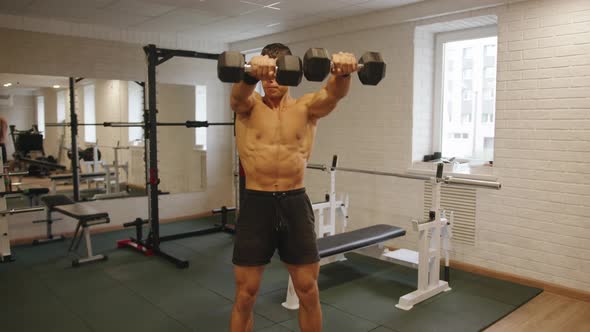 Young Muscular Man Training in the Gym and Raising Straight Arms with Dumbbells in Front of Him to