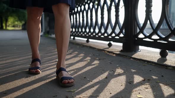 Female Feet in Black Sandals Walk on the Sandy Road Along the Openwork Lattice