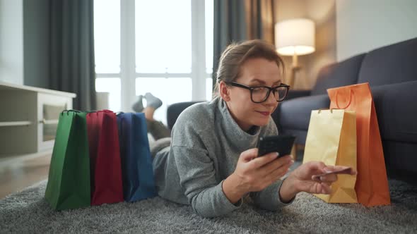 Happy Woman with Glasses Is Lying on the Floor and Makes an Online Purchase Using a Credit Card