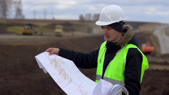 The Worker Looks at the Drawings on the Background of the Road Under Construction.