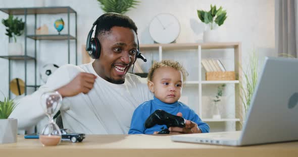 Black-Skinned Dad Playing Videogames on Laptop Together with Small Son and Celebrating Victory