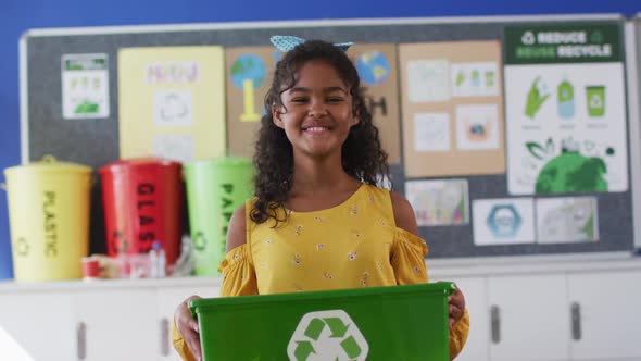 Mixed race schoolgirl smiling, holding recycling bin, standing in classroom