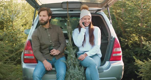 Woman which Sitting Together with Her Husband in Car's trunk Among Beautiful Fir Trees