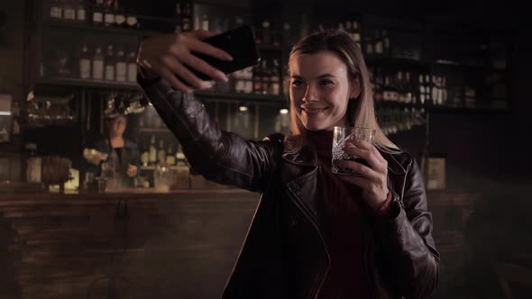 Young Woman Takes Selfie on Smartphone Holds Drink in Her Hand in Fashionable Bar Background of