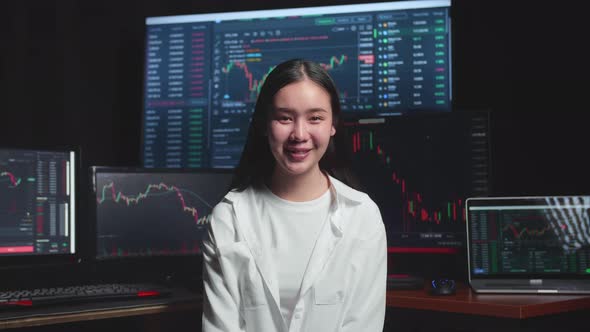 Asian Female Stock Market Broker Working On Computer And Smiling To Camera With Analysing Graphs