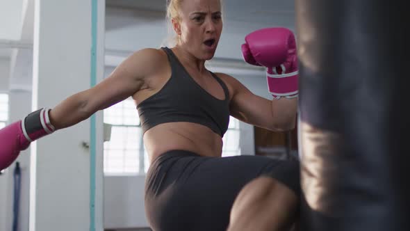 Caucasian female boxer wearing boxing gloves kicking the punching bag at the gym