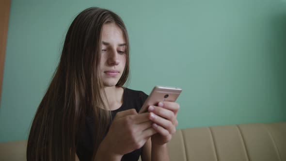 Attractive Blonde Caucasian Teen Girl Sitting on a Sofa at Home and Texting on Her Phone. Smiling