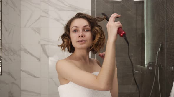 Portrait of a Young Woman in Big White Bath Towel Styling Her Hair with a Curling Iron in Front of a