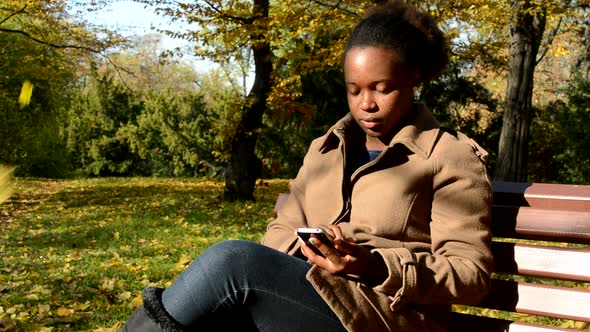 Young Beautiful African Serious Girl Sits on Bench in Woods and Works on Phone