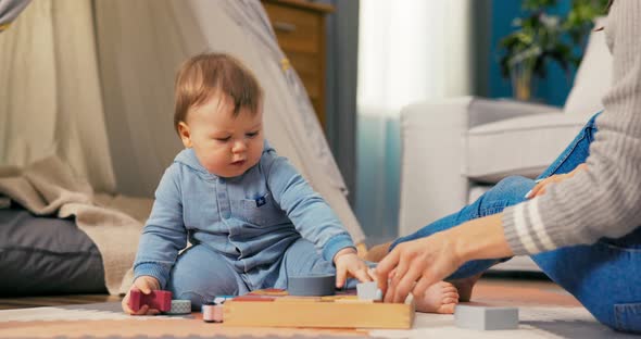 Small Sweet Child is Sitting on Floor Wearing Blue Bodysuit and Looking at Box Into