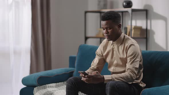 Young Afroamerican Man is Using Smartphone Sitting on Couch in Apartment Male Internet User Viewing