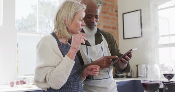 Mixed race senior couple wearing aprons using digital tablet while cooking in the kitchen at home