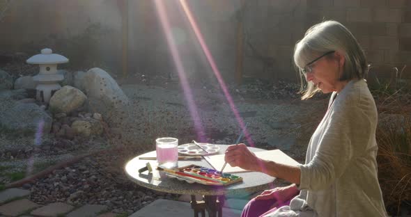 A beautiful old woman painting to relax and destress with watercolor in the sunlight of her outdoor