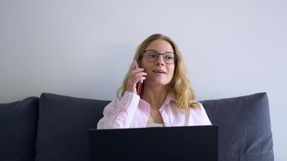 Beautiful Blonde Woman is Sitting at a Laptop at Home and Talking on the Phone with a Smile