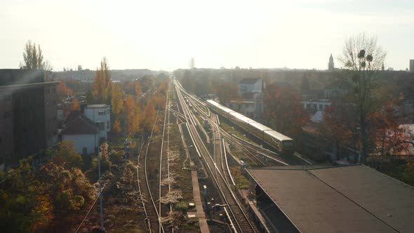 Scenic Aerial Shot of Passanger Train Driving Into Sunset on Train Tracks Surrounded By Treets Above