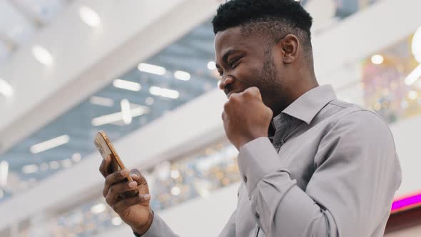 Young Happy Satisfied African American Guy Winner Looking at Phone Screen Celebrating Victory