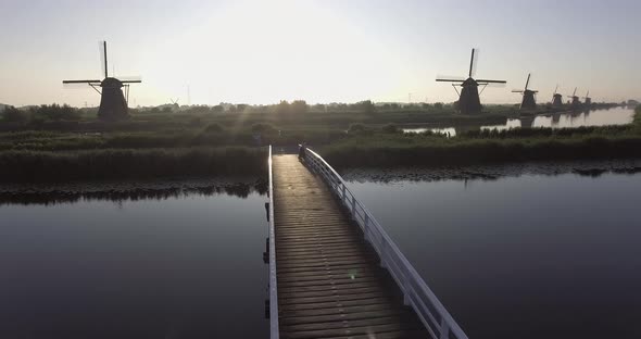 A drone shot panning left, around a girl standing on a bridge, looking at Dutch Windmills in the Net