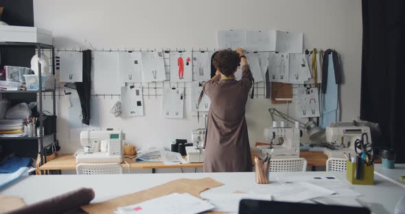 Female Clothes Designer Looking at Drawings of Garments Hanging on Wall