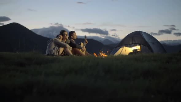 Group of Three Friends Play Guitar and Sing at Camp Fire in Nature Mountain Outdoor Camping Scene at