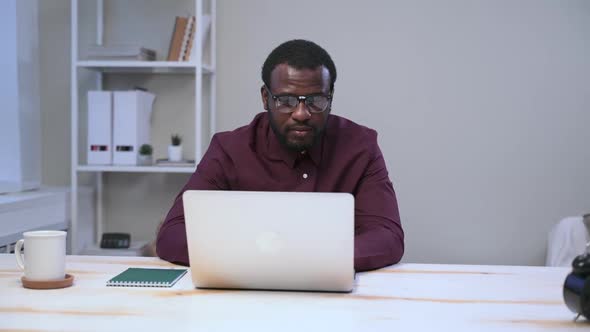 African Man in Glasses is Working on Laptop at Table and Smile Spbas