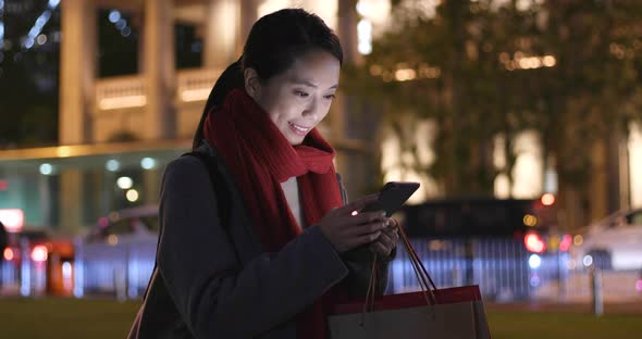 Woman use of smart phone in city at night and hold with shopping bag