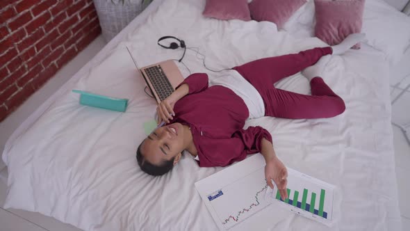 Young Cheerful Woman with Toothy Smile Lying on Bed with Business Graphs and Laptop Talking on