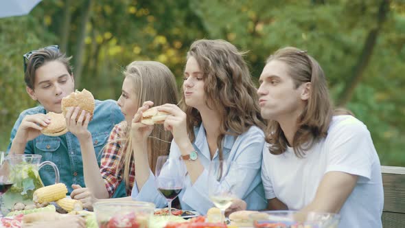 Friends Eating Burgers Sitting At Dinner Table At Outdoor Party