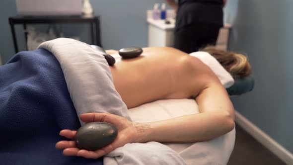 Masseuse or massage therapist uses heated stones on mature, beautiful woman’s back and hands. Steadi