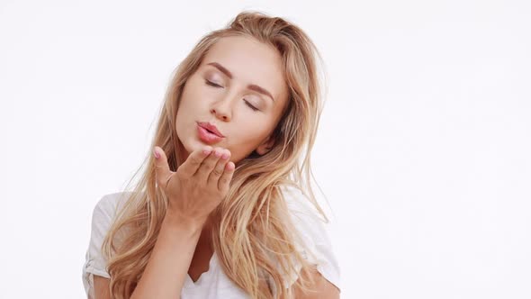 Young Beautiful Caucasian Blonde Girl Blowing a Kiss Picturing Imaginary Heart Smiling Shy on White