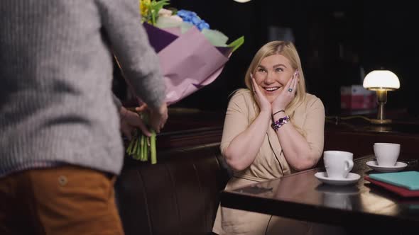 Portrait of Surprised Excited Woman Sitting in Restaurant As Boy Giving Bouquet of Flowers in Slow