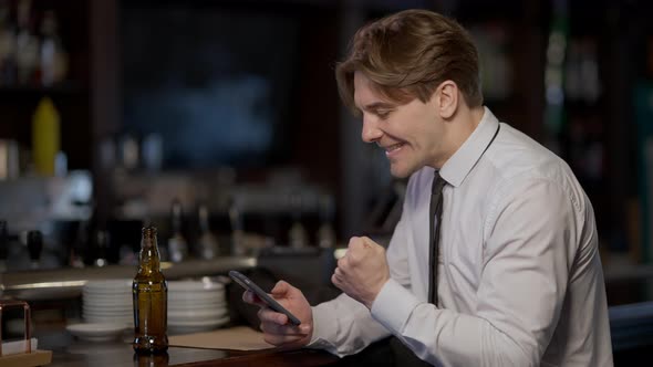 Joyful Adult Caucasian Man Making Victory Gesture Gaming Gambling Online on Smartphone Drinking Beer