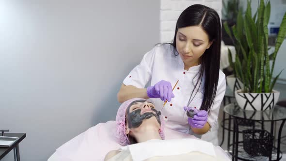 Pretty cosmetologist with long hair makes a mask to a woman for cleansing the pores on the face